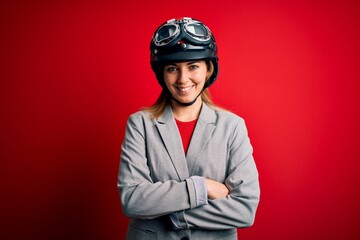 Young beautiful blonde motorcyclist woman wearing motorcycle helmet over red background happy face smiling with crossed arms looking at the camera. Positive person.