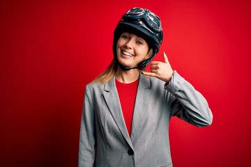 Young beautiful blonde motorcyclist woman wearing motorcycle helmet over red background smiling doing phone gesture with hand and fingers like talking on the telephone. Communicating concepts.
