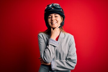 Young beautiful blonde motorcyclist woman wearing motorcycle helmet over red background looking confident at the camera smiling with crossed arms and hand raised on chin. Thinking positive.