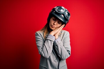 Young beautiful blonde motorcyclist woman wearing motorcycle helmet over red background sleeping tired dreaming and posing with hands together while smiling with closed eyes.