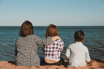 children sit on the sand and look at the water. View from the back. Two girls and a boy look at the...