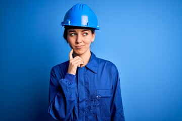 Young beautiful worker woman with blue eyes wearing security helmet and uniform with hand on chin thinking about question, pensive expression. Smiling with thoughtful face. Doubt concept.