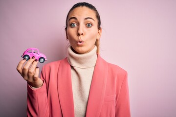 Young beautiful blonde woman with blue eyes holding small pink car over isolated background scared in shock with a surprise face, afraid and excited with fear expression