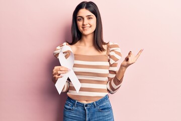 Young beautiful girl holding white cancer ribbon smiling happy pointing with hand and finger to the side