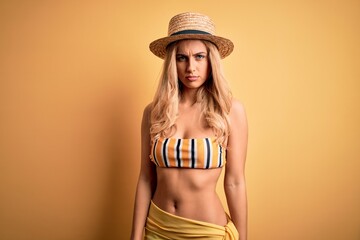 Young beautiful blonde woman on vacation wearing bikini and hat over yellow background skeptic and nervous, frowning upset because of problem. Negative person.