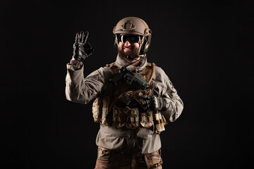 Naklejka premium USA soldier in a military suit with a rifle smiles and shows the okay sign against a dark background, an American commando in uniform
