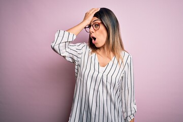 Young beautiful woman wearing casual striped t-shirt and glasses over pink background surprised...