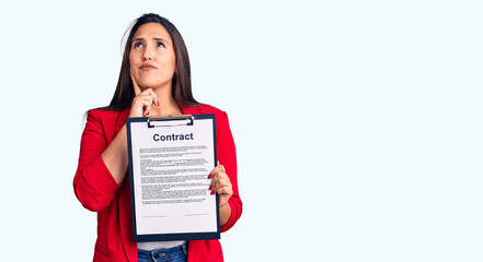 Young beautiful brunette woman holding clipboard with contract serious face thinking about question...