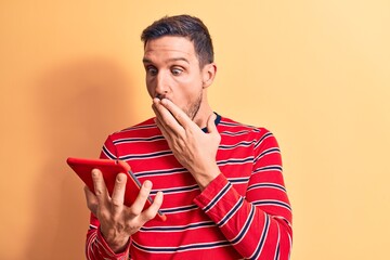 Young handsome man holding touchpad standing over isolated yellow background covering mouth with...