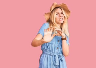 Young beautiful blonde woman wearing summer hat and dress disgusted expression, displeased and...