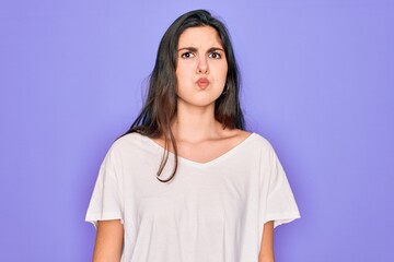 Fototapeta premium Young beautiful brunette woman wearing casual white t-shirt over purple background puffing cheeks with funny face. Mouth inflated with air, crazy expression.