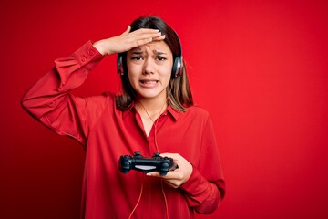 Young beautiful brunette gamer girl playing video game using joystick and headphones stressed with hand on head, shocked with shame and surprise face, angry and frustrated. Fear and upset for mistake. © Krakenimages.com
