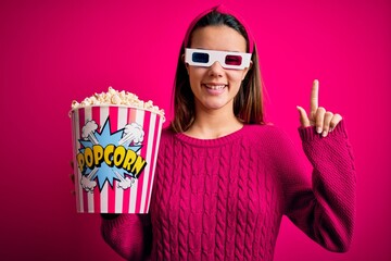 Obraz premium Young beautiful girl watching movie using 3d glasses eating box with popcorns with a big smile on face, pointing with hand finger to the side looking at the camera.