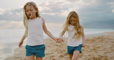 Happy young twin girls walking down the beach playing at sunset, summer family lifestyle - Powered by Adobe