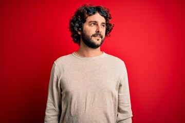 Young handsome man with beard wearing casual sweater standing over red background smiling looking to the side and staring away thinking.