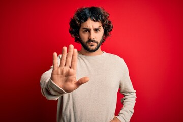 Young handsome man with beard wearing casual sweater standing over red background doing stop sing...