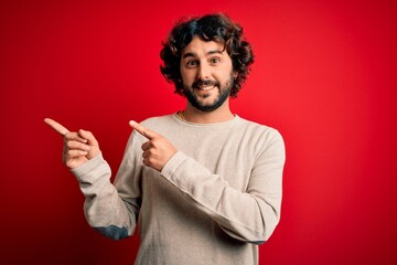 Young handsome man with beard wearing casual sweater standing over red background smiling and looking at the camera pointing with two hands and fingers to the side.