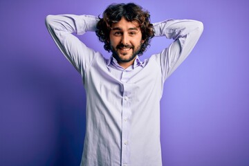 Naklejka premium Young handsome business man with beard wearing shirt standing over purple background relaxing and stretching, arms and hands behind head and neck smiling happy