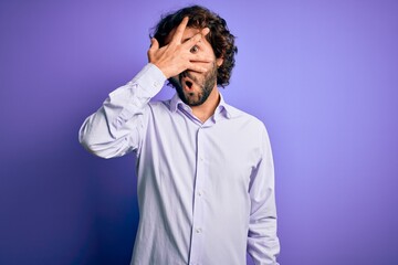 Fototapeta premium Young handsome business man with beard wearing shirt standing over purple background peeking in shock covering face and eyes with hand, looking through fingers with embarrassed expression.