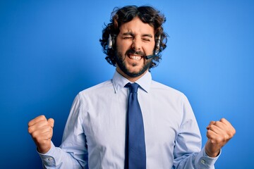 Young handsome call center agent man with beard working using headset over blue background very happy and excited doing winner gesture with arms raised, smiling and screaming for success. Celebration