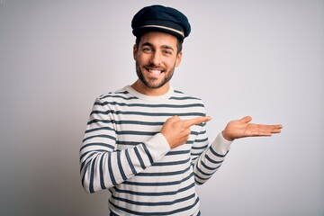 Young handsome sailor man with beard wearing navy striped uniform and captain hat amazed and smiling to the camera while presenting with hand and pointing with finger.