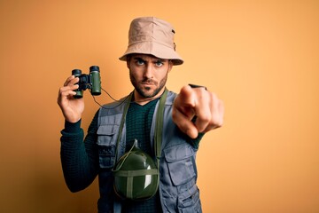 Young handsome tourist man with beard on vacation wearing explorer hat using binoculars pointing with finger to the camera and to you, hand sign, positive and confident gesture from the front