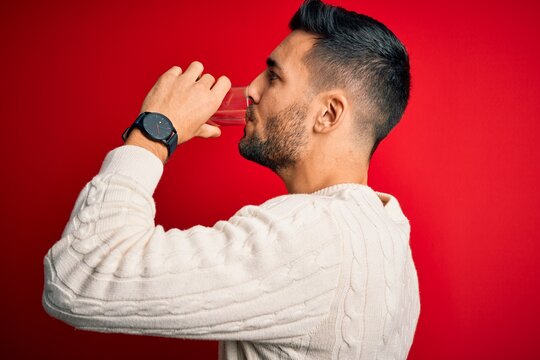 Young handsome man drinking glass of healthy water to refreshment standing over isolated red background