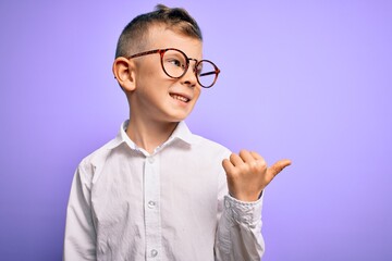 Young little caucasian kid with blue eyes wearing glasses and white shirt over purple background smiling with happy face looking and pointing to the side with thumb up.