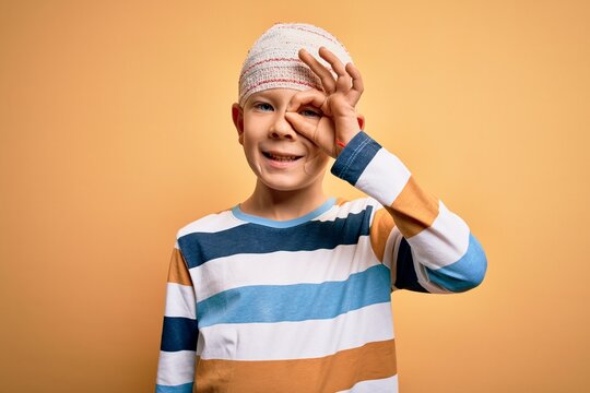 Young little caucasian kid injured wearing medical bandage on head over yellow background doing ok gesture with hand smiling, eye looking through fingers with happy face.