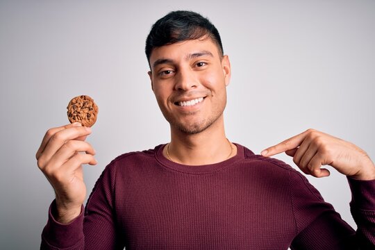 Young hispanic man eating chocolate chips cookie over isolated background with surprise face pointing finger to himself