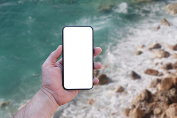 Guy holds in his hand a smartphone close-up, with a white screen on a background of sea. Mock-up Technology.