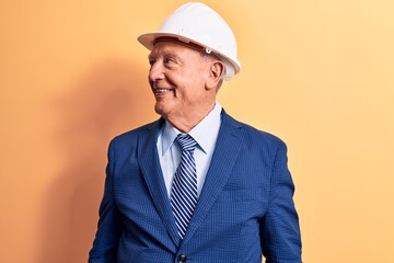 Senior grey-haired architect man wearing suit and security hardhat over yellow background looking to side, relax profile pose with natural face and confident smile.