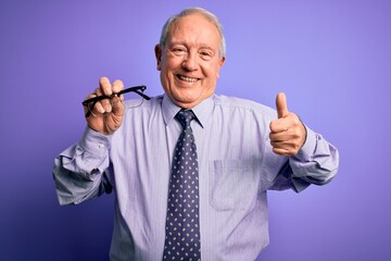 Senior grey haired man holding correction glasses over purple background happy with big smile doing ok sign, thumb up with fingers, excellent sign