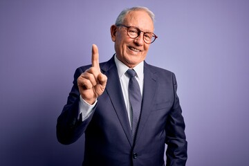 Grey haired senior business man wearing glasses and elegant suit and tie over purple background pointing finger up with successful idea. Exited and happy. Number one.