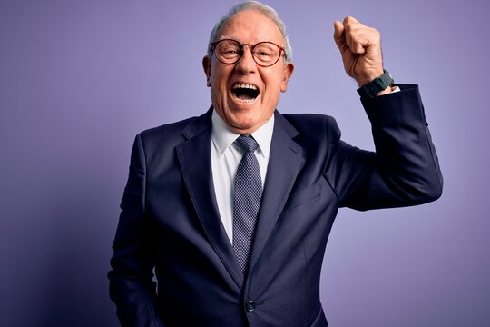 Grey haired senior business man wearing glasses and elegant suit and tie over purple background angry and mad raising fist frustrated and furious while shouting with anger. Rage and aggressive