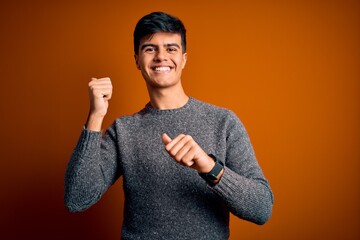 Young handsome man wearing casual sweater standing over isolated orange background Pointing to the back behind with hand and thumbs up, smiling confident