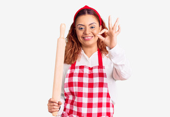 Young latin woman wearing apron holding kneader doing ok sign with fingers, smiling friendly gesturing excellent symbol