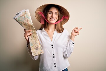 Young beautiful redhead woman wearing asian traditional hat holding city map very happy pointing with hand and finger to the side