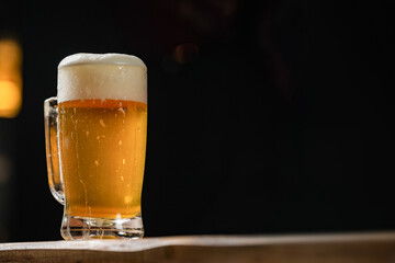 cold mug with beer, with overflowing foam, on wooden table and dark background