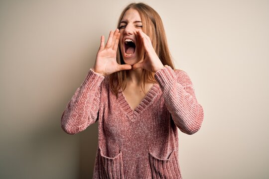 Young beautiful redhead woman wearing pink casual sweater over isolated white background Shouting angry out loud with hands over mouth