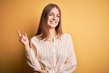 Young beautiful redhead woman wearing casual striped shirt over isolated yellow background with a big smile on face, pointing with hand and finger to the side looking at the camera.