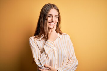 Young beautiful redhead woman wearing casual striped shirt over isolated yellow background looking confident at the camera with smile with crossed arms and hand raised on chin. Thinking positive.