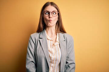 Young beautiful redhead woman wearing jacket and glasses over isolated yellow background making fish face with lips, crazy and comical gesture. Funny expression.