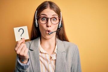 Young redhead call center agent woman using headset holding question mark reminder scared in shock...