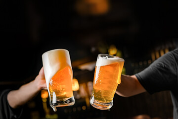 two hands with beer mugs, toasting in celebration, with overflowing foam, dark background and space for writing - obrazy, fototapety, plakaty