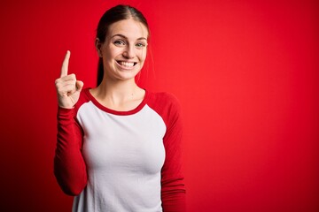 Young beautiful redhead woman wearing casual t-shirt over isolated red background pointing finger up with successful idea. Exited and happy. Number one.