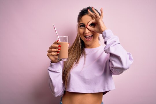 Young beautiful fitness woman drinking protein chocolate milkshake before gym with happy face smiling doing ok sign with hand on eye looking through fingers