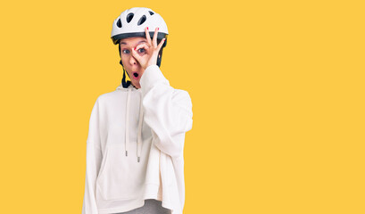 Beautiful brunette young woman wearing bike helmet and sporty clothes doing ok gesture shocked with surprised face, eye looking through fingers. unbelieving expression.