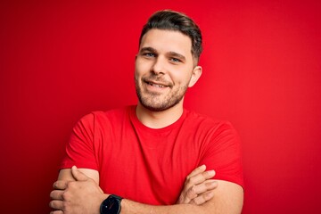 Young man with blue eyes wearing casual t-shirt over red isolated background happy face smiling with crossed arms looking at the camera. Positive person.