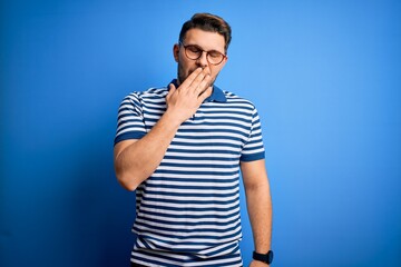 Young man with blue eyes wearing glasses and casual striped t-shirt over blue background bored yawning tired covering mouth with hand. Restless and sleepiness.